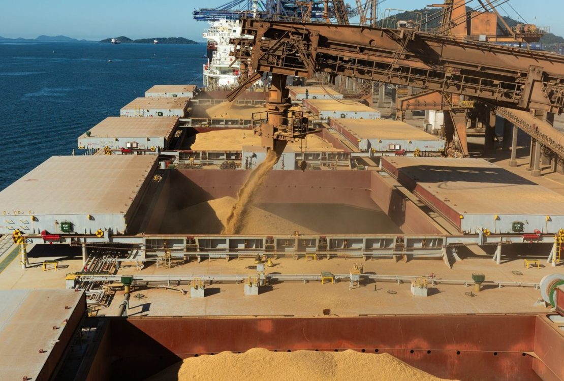 Vista aérea de um navio cargueiro sendo carregado com grãos em um terminal portuário industrial.