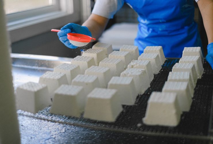 Trabalhador polvilha ingredientes sobre blocos de queijo fresco em uma unidade de produção de laticínios.