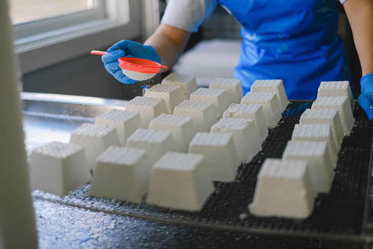 Trabalhador polvilha ingredientes sobre blocos de queijo fresco em uma unidade de produção de laticínios.
